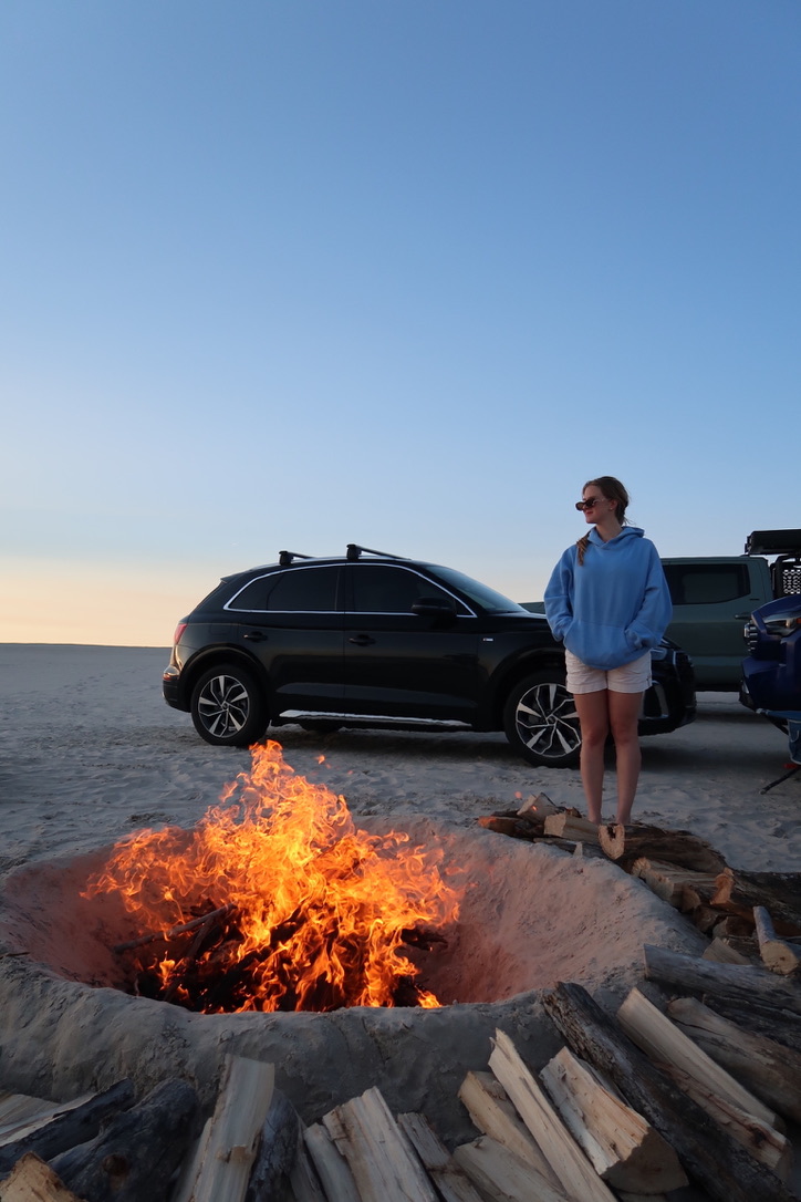 Shelby beside a beach bonfire with her car at sunrise